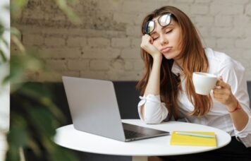 Portrait of a tired young businesswoman sitting at the table with laptop computer while holding cup of coffee and sleeping at a cafe